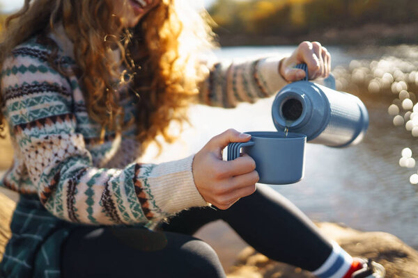 Beautiful woman traveler in a hat sits on a log near the river, drinks a hot drink from a thermos. Smiling female tourist enjoys the autumn landscape near the river. The concept of travel, relax.