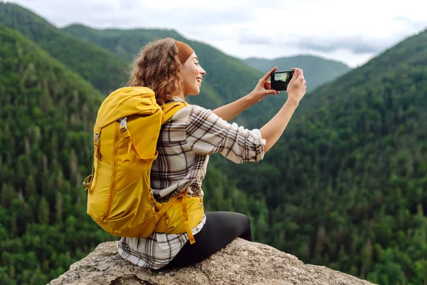 stock image Young traveler with a yellow backpack is resting and taking pictures on the phone on a mountain cliff, admiring the landscape of the high Tatras, mountain range. Beautiful woman on top of the mountain