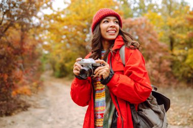 Sonbahar ormanında yürüyüş yaparken elinde fotoğraf makinesi ve şık bir kazakla gezen mutlu bir bayan turist. Bir kadın fotoğraf çeker ve doğanın güzelliğinden zevk alır..