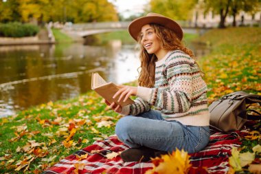 Şapkalı ve şık kazaklı güzel bir kadın sonbahar parkında bir paspasın üzerinde oturur ve kitap okur. Genç turistler doğayla baş başa kalmanın ve havanın tadını çıkarıyorlar..