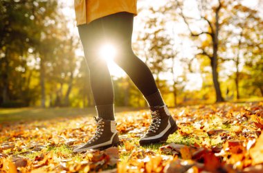 Güneşli bir sonbahar parkında yaprakların arasında dağlık bir alanda yürüyüş botlarıyla ayakların yakın çekimi. Yaşam tarzı konsepti, doğa yürüyüşü..