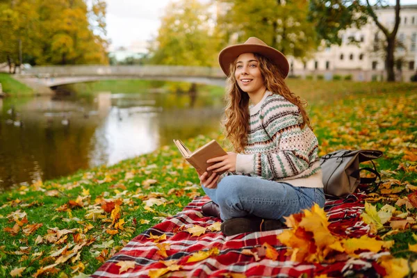 Şapkalı ve şık kazaklı güzel bir kadın sonbahar parkında bir paspasın üzerinde oturur ve kitap okur. Genç turistler doğayla baş başa kalmanın ve havanın tadını çıkarıyorlar..