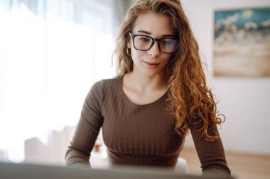 Genç bir kadın evde dizüstü bilgisayar kullanıyor, ekrana bakıyor, masada okuyor ya da yazıyor. Kadın evde dizüstü bilgisayarla çalışıyor. Serbest çalışma, tatil, eğitim kavramı.