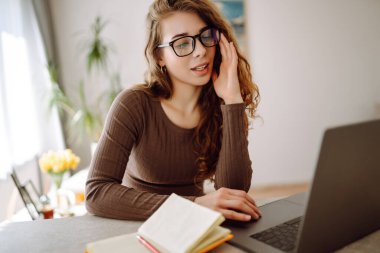 Genç bir kadın evde dizüstü bilgisayar kullanıyor, ekrana bakıyor, masada okuyor ya da yazıyor. Kadın evde dizüstü bilgisayarla çalışıyor. Serbest çalışma, tatil, eğitim kavramı.