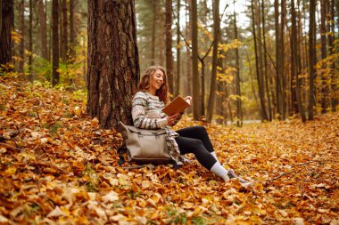 Kıvırcık saçlı ve elinde kitap olan bir kadın sonbahar ormanında bir akçaağaç ağacının altında oturur, okur ve havanın tadını çıkarır. Sonbahar, tatil kavramı.