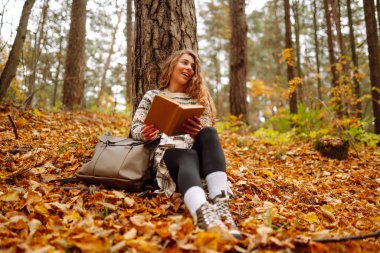 Kıvırcık saçlı ve elinde kitap olan bir kadın sonbahar ormanında bir akçaağaç ağacının altında oturur, okur ve havanın tadını çıkarır. Sonbahar, tatil kavramı.