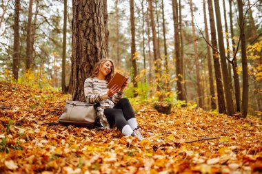 Kıvırcık saçlı ve elinde kitap olan bir kadın sonbahar ormanında bir akçaağaç ağacının altında oturur, okur ve havanın tadını çıkarır. Sonbahar, tatil kavramı.