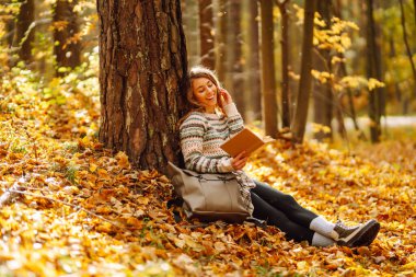Kıvırcık saçlı ve elinde kitap olan bir kadın sonbahar ormanında bir akçaağaç ağacının altında oturur, okur ve havanın tadını çıkarır. Sonbahar, tatil kavramı.