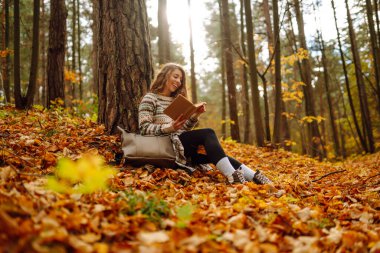Kıvırcık saçlı ve elinde kitap olan bir kadın sonbahar ormanında bir akçaağaç ağacının altında oturur, okur ve havanın tadını çıkarır. Sonbahar, tatil kavramı.