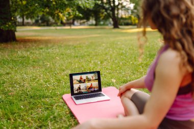 Spor giyimli atletik genç bir kadın laptopun önünde yoga minderinde oturup spor yapıyor. Çevrimiçi fitness derslerinde kadın eğitimi, gün batımında park çimlerinde webcam üzerinden spor eğitmeniyle konuşmak..