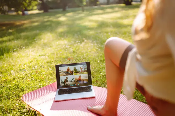 Athletic young woman in sportswear sits in front of laptop on yoga mat and works out. Woman training in online fitness classes, talking to sports trainer via webcam on park lawn at sunset.