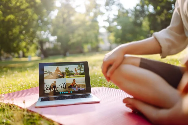 Athletic young woman in sportswear sits in front of laptop on yoga mat and works out. Woman training in online fitness classes, talking to sports trainer via webcam on park lawn at sunset.