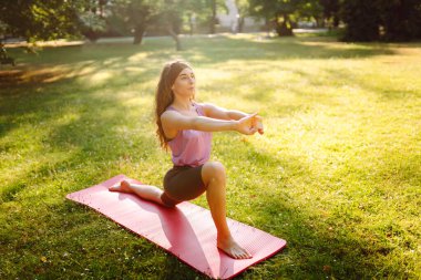 Spor salonundaki kadın yoga yapıyor, parkta spor yapıyor. Güneşli bir çimenlikte paspasın üzerinde oturan atletik bir kadın. Fitness. Etkin yaşam biçimi.