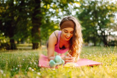 Spor salonundaki kadın yoga yapıyor, parkta spor yapıyor. Güneşli bir çimenlikte paspasın üzerinde oturan atletik bir kadın. Fitness. Etkin yaşam biçimi.