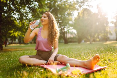 Spor salonundaki kadın yoga yapıyor, parkta spor yapıyor. Güneşli bir çimenlikte paspasın üzerinde oturan atletik bir kadın. Fitness. Etkin yaşam biçimi.