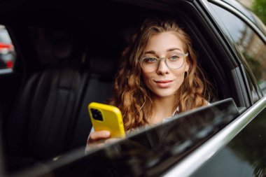 Güzel genç bir kadın arabanın arka koltuğunda otururken akıllı telefon kullanır. Teknoloji kavramı, araba ve iş seyahati..