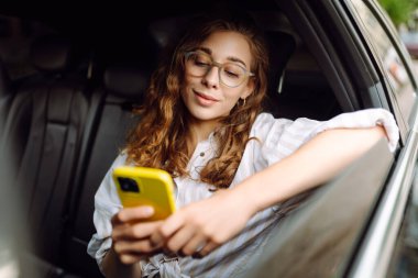 Güzel genç bir kadın arabanın arka koltuğunda otururken akıllı telefon kullanır. Teknoloji kavramı, araba ve iş seyahati..
