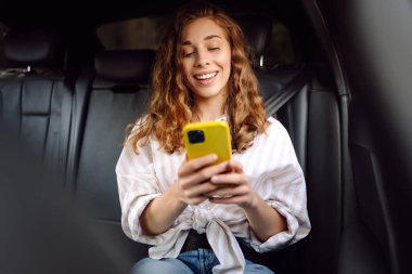 Güzel genç bir kadın arabanın arka koltuğunda otururken akıllı telefon kullanır. Teknoloji kavramı, araba ve iş seyahati..
