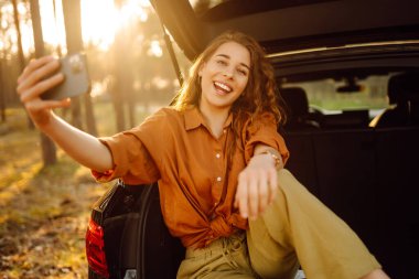 Bir arabanın bagajında telefonlu mutlu, şık bir bayan turist, selfie çekiyor, video görüşmesinde konuşuyor. Gün batımında arabanın bagajında bir kadın. Arabayla seyahat etmek, kamp yapmak. Etkin yaşam biçimi.