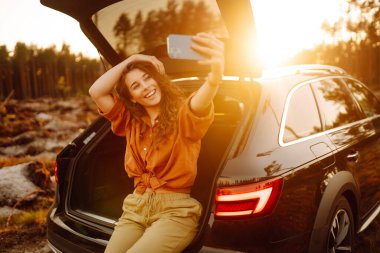 Bir arabanın bagajında telefonlu mutlu, şık bir bayan turist, selfie çekiyor, video görüşmesinde konuşuyor. Gün batımında arabanın bagajında bir kadın. Arabayla seyahat etmek, kamp yapmak. Etkin yaşam biçimi.