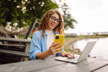 Parktaki bir masada dizüstü bilgisayar ve akıllı telefon kullanarak bir bankta oturan güzel bir bayan serbest yazar. Şehir yaşam tarzı. İş, blog yazma, serbest çalışma.
