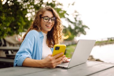 Parktaki bir masada dizüstü bilgisayar ve akıllı telefon kullanarak bir bankta oturan güzel bir bayan serbest yazar. Şehir yaşam tarzı. İş, blog yazma, serbest çalışma.