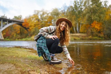 Süslü bir kazak ve sırt çantalı bir şapka giyen bir turist nehir manzarasına hayran. Şapkalı genç bir kız doğanın tadını çıkarıyor. Seyahat konsepti. Sonbahar havası.