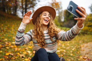 Elinde telefon olan şık bir süveter giyen güzel bir kadın, sonbahar parkındaki sarı yaprakların arasında bir açıklıkta oturuyor. Turistler havanın tadını çıkarır, sarı yapraklı bir fotoğraf çeker.. 
