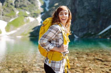 Dağların arasında turkuaz bir gölün arka planında sarı bir sırt çantasıyla gülümseyen bir bayan gezgin. Aktif yaşam tarzı. Yürüyüş, macera konsepti.
