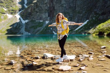Dağların arasında turkuaz bir gölün arka planında sarı bir sırt çantasıyla gülümseyen bir bayan gezgin. Aktif yaşam tarzı. Yürüyüş, macera konsepti.