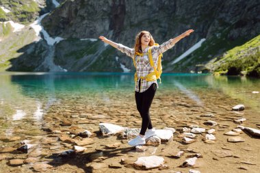 Dağların arasında turkuaz bir gölün arka planında sarı bir sırt çantasıyla gülümseyen bir bayan gezgin. Aktif yaşam tarzı. Yürüyüş, macera konsepti.