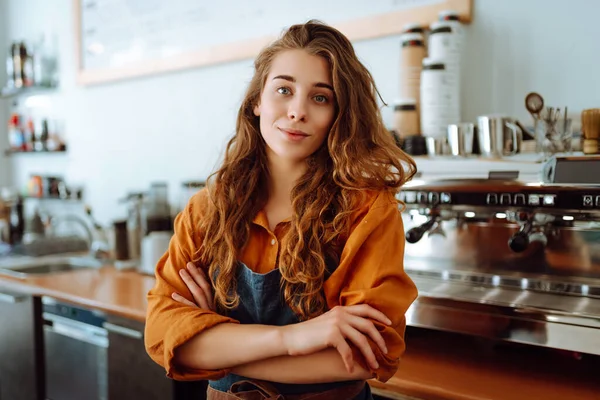 Portrait of a happy female barista standing behind the counter in a coffee shop. A woman cafe owner in an apron looks at the camera and smiles. Business concept.