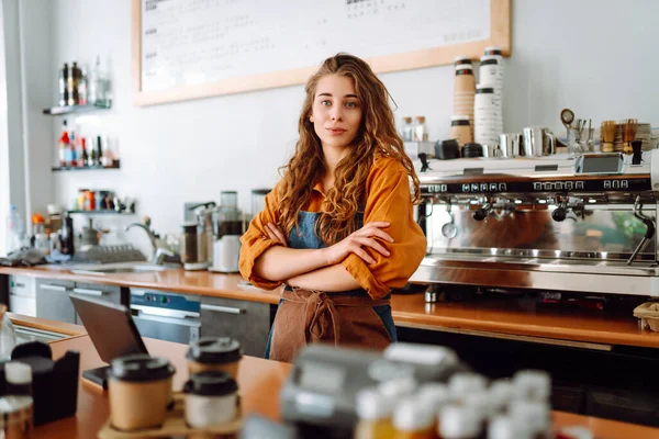 Portrait of a happy female barista standing behind the counter in a coffee shop. A woman cafe owner in an apron looks at the camera and smiles. Business concept.