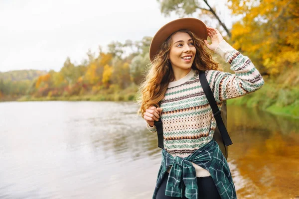 Süslü bir kazak ve sırt çantalı bir şapka giyen bir turist nehir manzarasına hayran. Şapkalı genç bir kız doğanın tadını çıkarıyor. Seyahat konsepti. Sonbahar havası.
