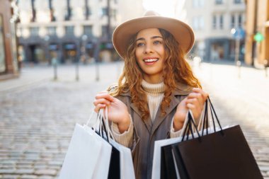 Dükkânın yanında alışveriş yaptıktan sonra şapkalı şık bir kadının portresi. Alışveriş torbalı kıvırcık saçlı kadın güneşli bir şehirde yürüyor. Satış kavramı, alışveriş.