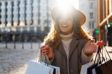 Dükkânın yanında alışveriş yaptıktan sonra şapkalı şık bir kadının portresi. Alışveriş torbalı kıvırcık saçlı kadın güneşli bir şehirde yürüyor. Satış kavramı, alışveriş.