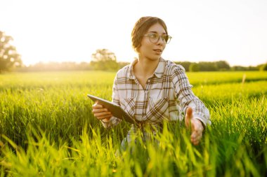 Modern tabletli bir kadın çiftçi filizleri eliyle değerlendirir, tarladaki yeşil filizleri. Tarım sektöründe dijital tabletle tarım.