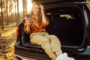 Günlük giysiler içinde genç bir bayan turist bir arabanın bagajında oturuyor ve telefon kullanıyor. Bir arabanın bagajında güzel bir kadın ormanda doğanın tadını çıkarıyor. Macera, teknoloji konsepti.
