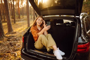 Günlük giysiler içinde genç bir bayan turist bir arabanın bagajında oturuyor ve telefon kullanıyor. Bir arabanın bagajında güzel bir kadın ormanda doğanın tadını çıkarıyor. Macera, teknoloji konsepti.