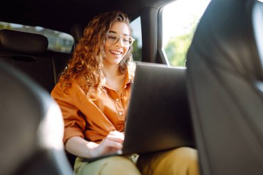 Modern bir dizüstü bilgisayarı olan genç bir iş kadını arabanın arka koltuğunda oturuyor. Başarılı bir başlangıç, uzak çalışma. Teknoloji kavramı, serbest çalışma..