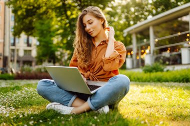 Genç bir kadın güneşli bir parkta oturuyor ve bir dizüstü bilgisayarla, blogla, çevrimiçi öğrenmeyle çalışıyor. Uzaktan çalışma, serbest çalışma, teknoloji kavramı.