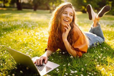 Genç bir kadın güneşli bir parkta oturuyor ve bir dizüstü bilgisayarla, blogla, çevrimiçi öğrenmeyle çalışıyor. Uzaktan çalışma, serbest çalışma, teknoloji kavramı.