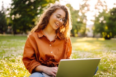 Genç bir kadın güneşli bir parkta oturuyor ve bir dizüstü bilgisayarla, blogla, çevrimiçi öğrenmeyle çalışıyor. Uzaktan çalışma, serbest çalışma, teknoloji kavramı.