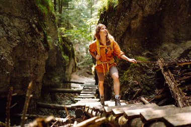 Sarı sırt çantalı, yürüyüş kıyafetli mutlu bir kadın dağlardaki ahşap yürüyüş yolu boyunca yürüyor. Yürüyüş, aktif yaşam tarzı. Açık hava macera konsepti.