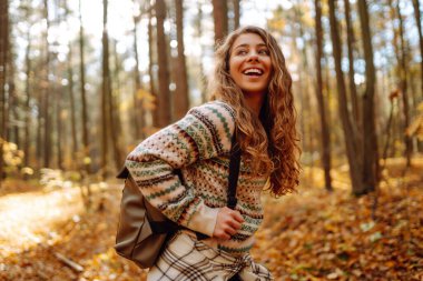 Sırt çantalı bir turist, sonbahar ormanında bir patika boyunca yürüyor. Güzel bir kadın doğada güneşli bir günün tadını çıkarır, özgürlüğü hisseder ve temiz havayı soluyarak doğayı keşfeder..