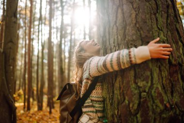 Sırt çantalı bir turist, sonbahar ormanında bir patika boyunca yürüyor. Güzel bir kadın doğada güneşli bir günün tadını çıkarır, özgürlüğü hisseder ve temiz havayı soluyarak doğayı keşfeder..