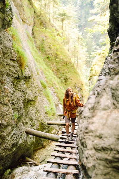 Happy woman in hiking clothes with a yellow backpack walks along a wooden hiking path in the mountains. Hiking, active lifestyle. Outdoor adventure concept.