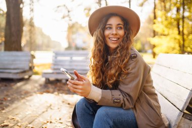 Bir sonbahar parkında bankta otururken şapkalı ve ceketli hoş bir bayan elinde bir akıllı telefon tutuyor. Tatil kavramı, teknoloji, hafta sonu.