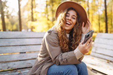 Bir sonbahar parkında bankta otururken şapkalı ve ceketli hoş bir bayan elinde bir akıllı telefon tutuyor. Tatil kavramı, teknoloji, hafta sonu.