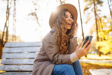 Bir sonbahar parkında bankta otururken şapkalı ve ceketli hoş bir bayan elinde bir akıllı telefon tutuyor. Tatil kavramı, teknoloji, hafta sonu.